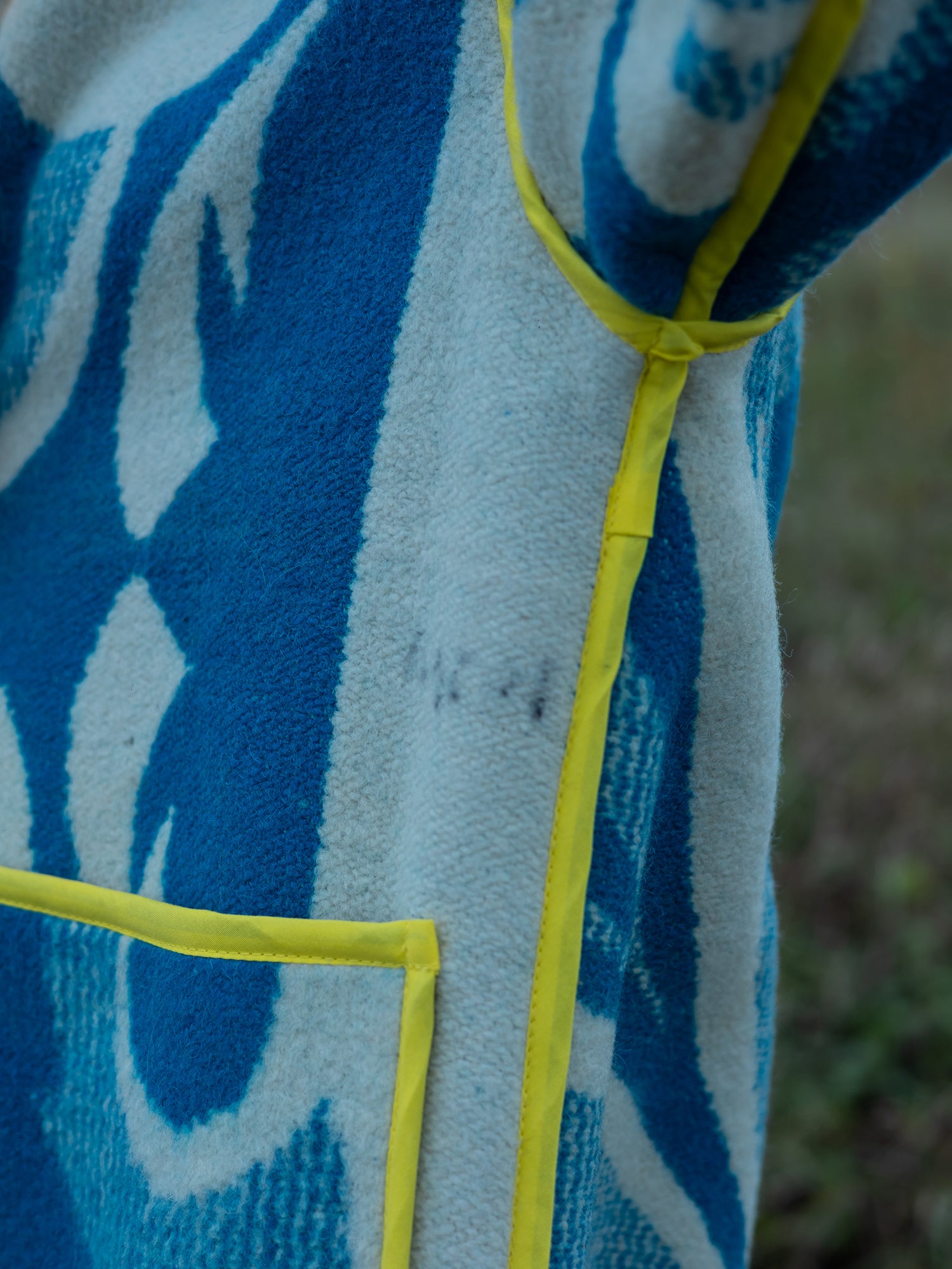 Close-up of a blue and white patterned upcycled blanket coat with yellow trim on a grassy background