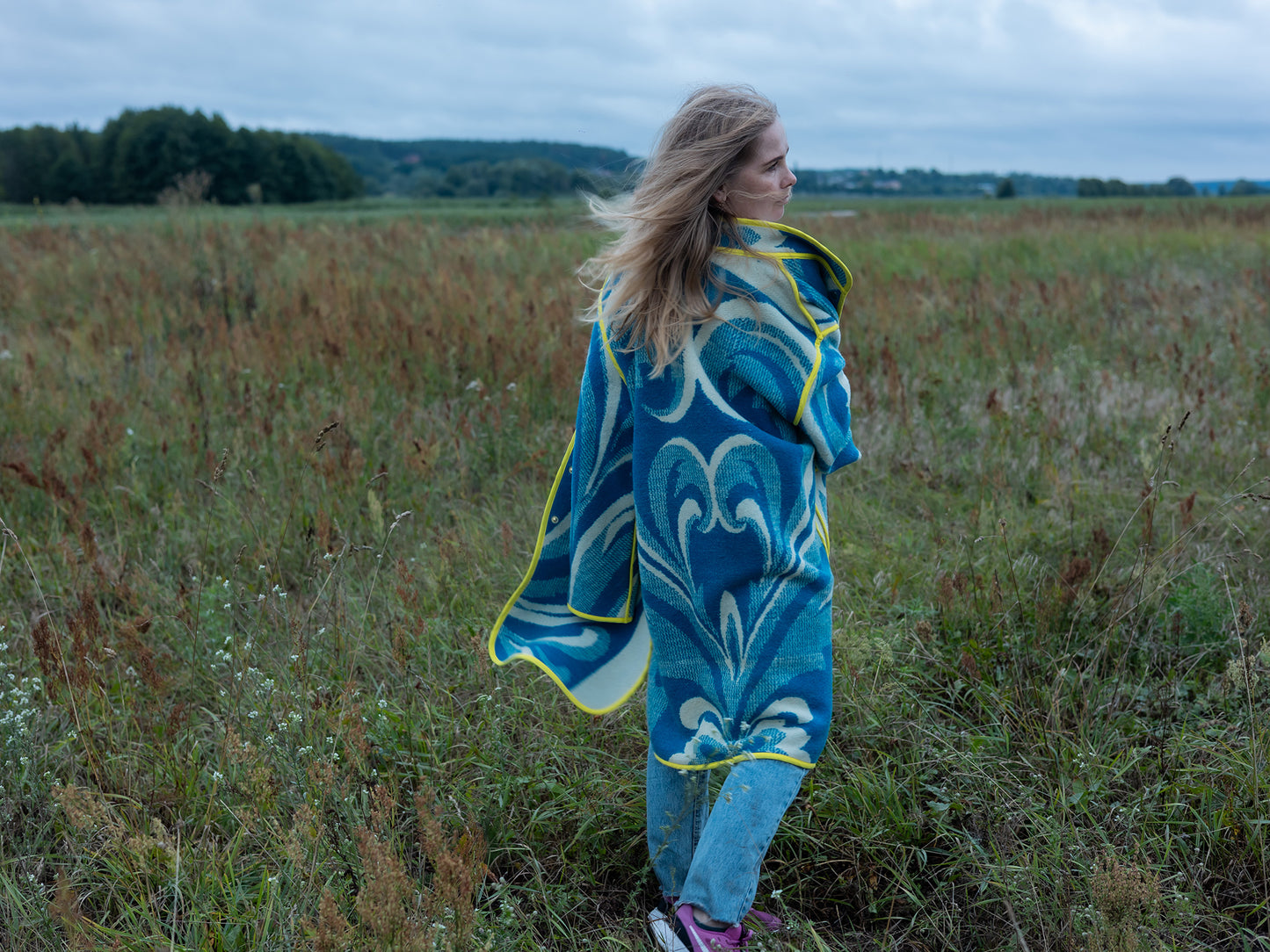 Woman wearing a blue patterned upcycled blanket coat standing in a field on a cloudy day